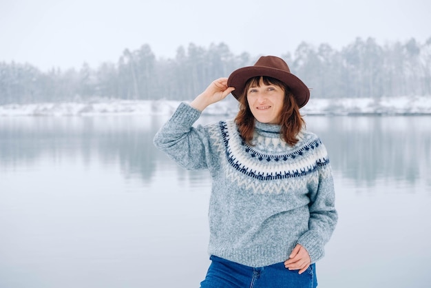 Woman in a brown hat and sweater stands on the shores of a snowcovered lake and forest