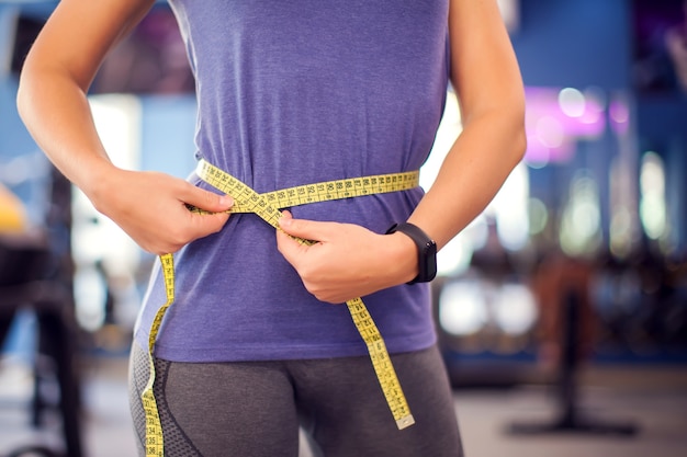 Woman in bue shirt measuring waist with meter in the gym. People, fitness and health concept