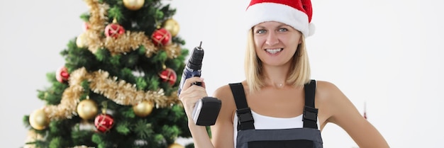 Woman builder in red santa claus hat holding drill in hands near christmas tree corporate new