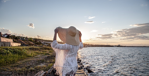 Woman by the sea at sunset back view. Dressed in a long skirt and hat.