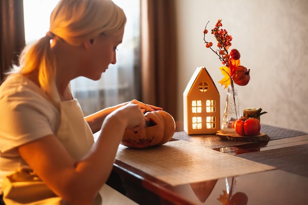 A woman carves a pumpkin for Halloween in a room with autumn decor and a lamp house. Cosy home and preparing for Halloween