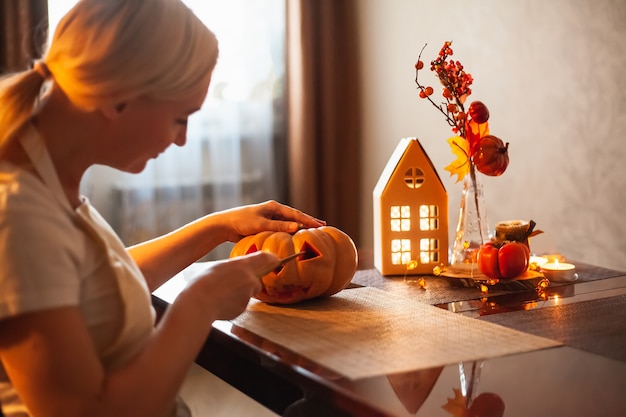 A woman carves a pumpkin for Halloween in a room with autumn decor and a lamp house. Cosy home and preparing for Halloween