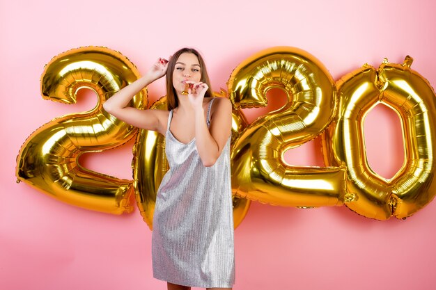 Woman celebrating new year blowing christmas pipe with 2020 balloons isolated over pink