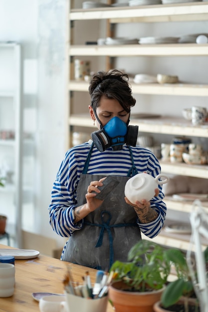 Woman ceramist wearing respirator polishing pottery in workshop female potter at workplace