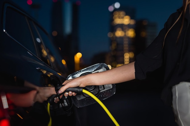 Woman charging a black EV car at a downtown charging station at night Renewable energy concept