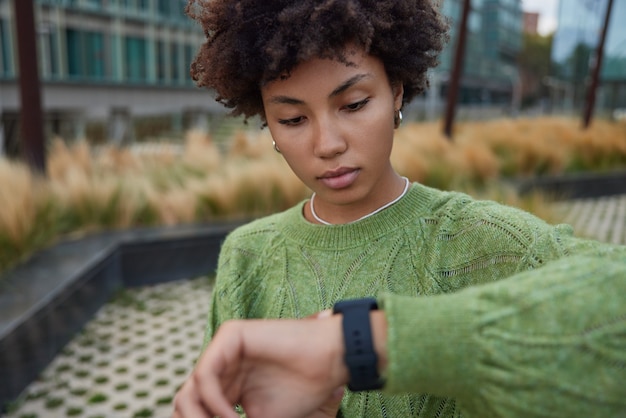 woman checks time on watch has appointment monitors covered distance on smartwatch after walking in city wears casual green jumper poses outdoors