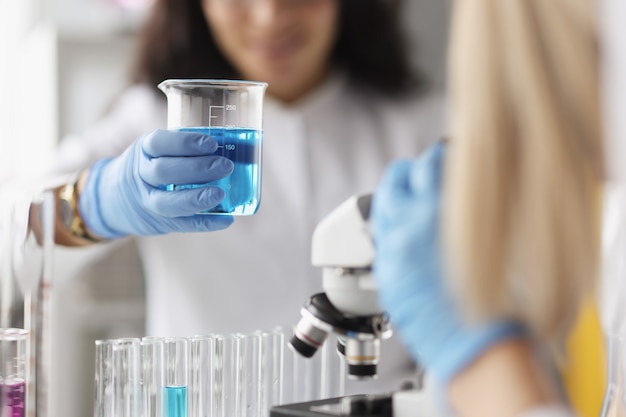 Woman chemist holding glass beaker with blue liquid in front of microscope in laboratory