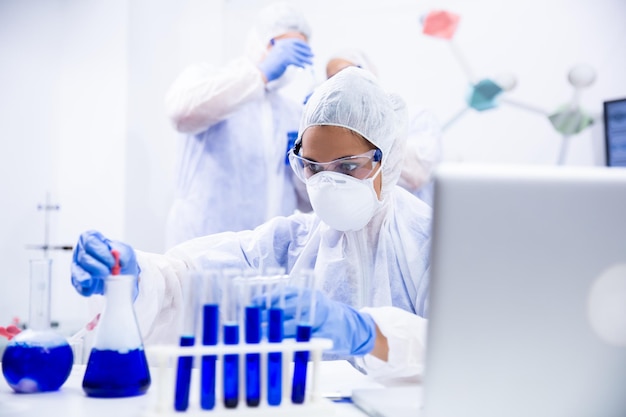 Woman chemist who works at the table and has 2 colleagues in the back and in front of the tube and laptop