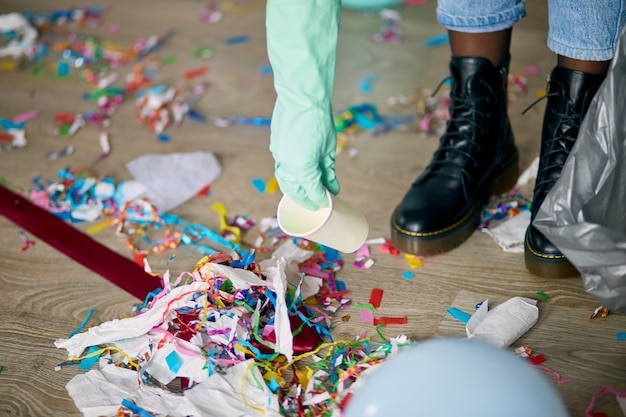 Woman cleaning mess of floor in room after party, removes garbage from the floor, disposable cups in a bag, morning after party celebration, housework, cleaning service