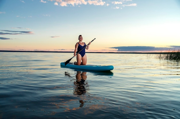 A woman in a closed swimsuit on her knees on a SUP board with an oar floats on the water against the background of the sunset sky