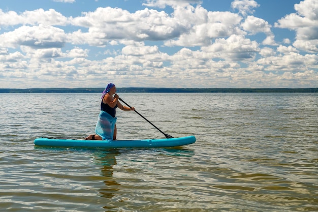 A woman in a closed swimsuit a pareo and a headdress on her knees on a SUP board with an oar floats on the water