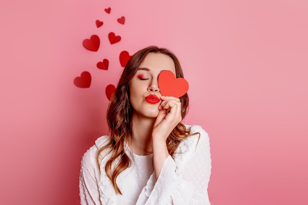 Woman closes one eye with a heart and sends an air kiss isolated on pink background