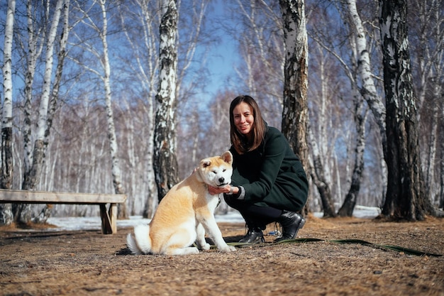 Woman in coat and an akita inu puppy in a winter birch park look at the camera