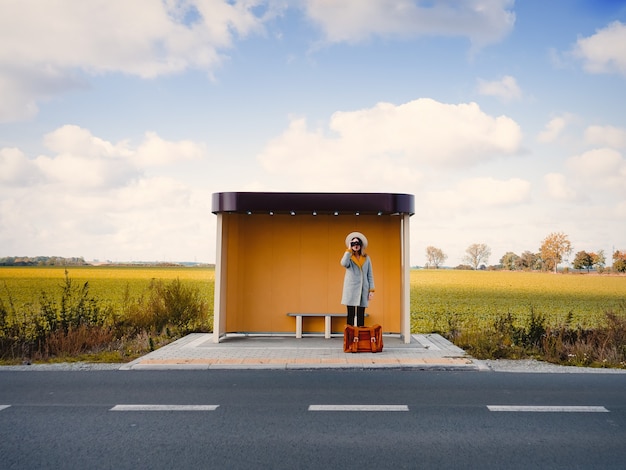 Woman in coat with suitcase and binocular on countryside bus stop