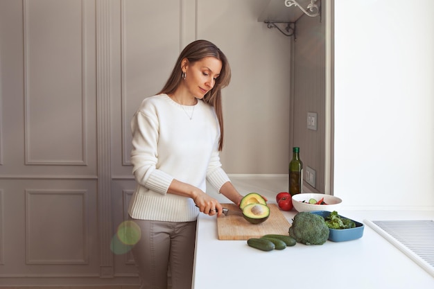 Woman cooking vegetarian salad with fresh vegetables. Model cutting with knife avocados, broccoli, kale salad, cucumbers and tomatoes in white kitchen