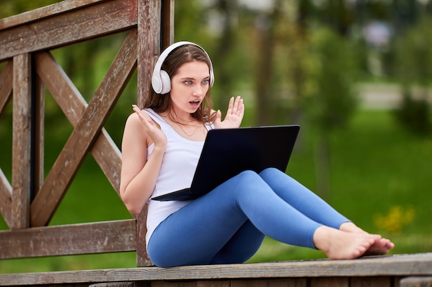 Woman in cordless headphones is surprising during online education with laptop in park