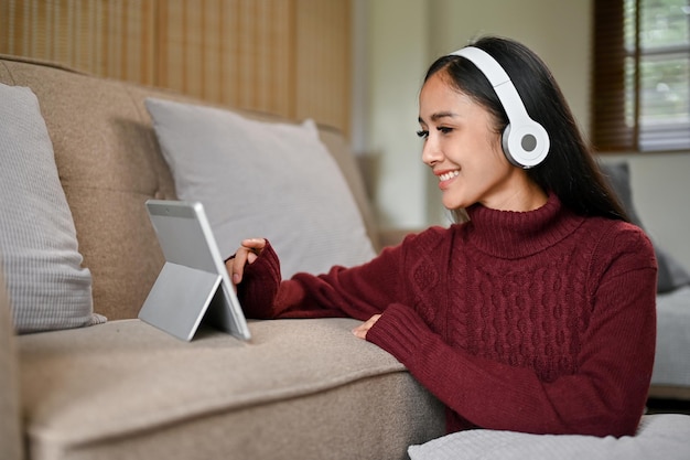 A woman in a cosy sweater wearing headphones and using her tablet on a sofa in her living room