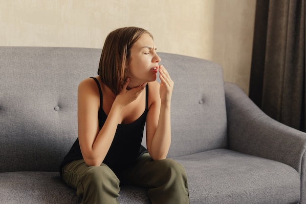 Woman coughs, covering her mouth with a hand, feeling cold and a sore throat, sitting on grey sofa, at home.