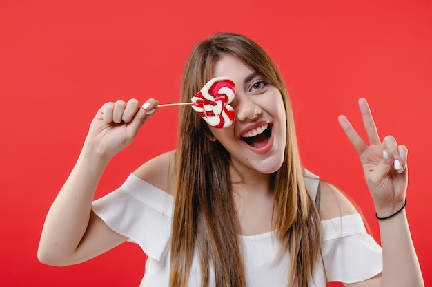 Photo woman covering eye with heart shaped lollypop candy wearing white blouse isolated on red wall