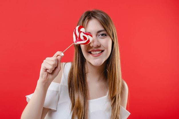 Woman covering eye with heart shaped lollypop candy wearing white blouse isolated on red wall