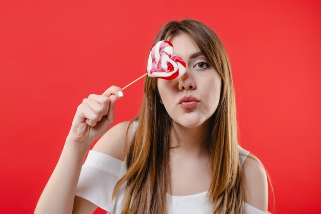 Woman covering eye with heart shaped lollypop candy wearing white blouse isolated on red wall