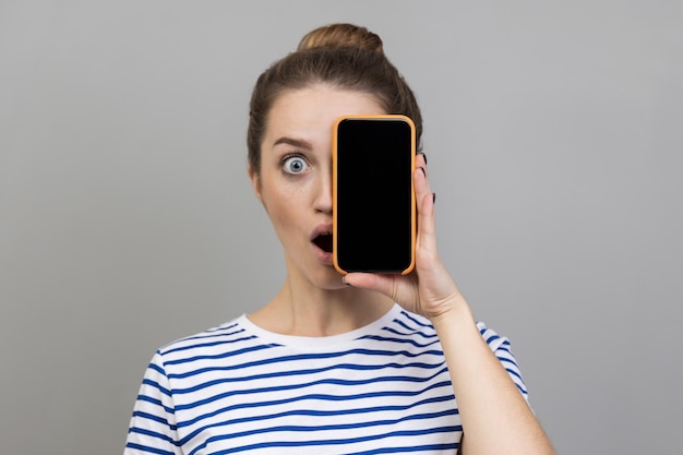 Woman covering half face with mobile phone and looking in surprise at camera technology and identity