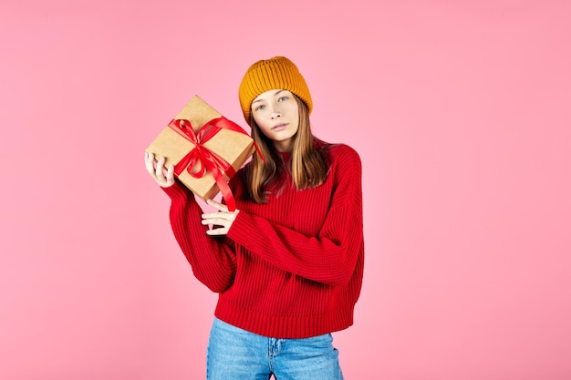 Photo woman in cozy red sweater holding gift boxes in hands and choosing present over pink background indecisive young hipster girl wearing knitted hat choosing a present box