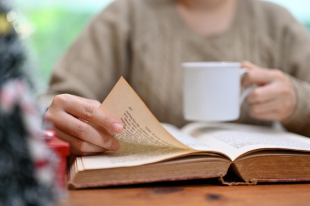 Woman in cozy sweater reading a book or novel while sipping hot coffee at her desk cropped