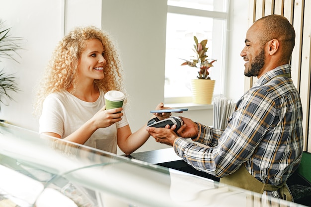 Woman customer of coffee shop paying for coffee through mobile phone using contactless technology