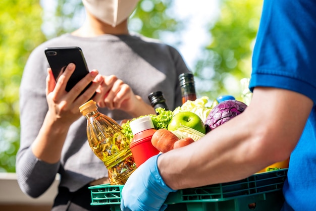 Woman customer wearing face mask checking grocery that ordered online and delivered by deliveryman at home, food delivery service in the times of COVID-19 concept