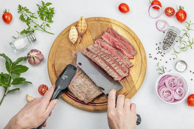 Woman cuts grilled steak Female hands with knife and culinary tong Fresh herbs spices vegetables Wooden cooking board picnic or barbecue concept Light stone concrete background top view