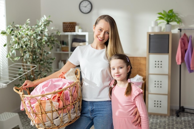 Woman and daughter embrace standing in background of laundry room bathroom mother holds wicker laundry basket stuffed with colorful clothes spending time together while doing household chores