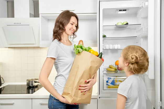 Woman and daughter with products bag near empty fridge.