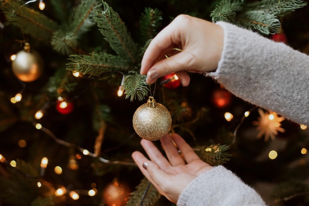 Woman decorating christmas tree with shiny golden bauble closeup Preparation for christmas time Modern glitter ornament in hands on background of festive tree in lights Happy holidays