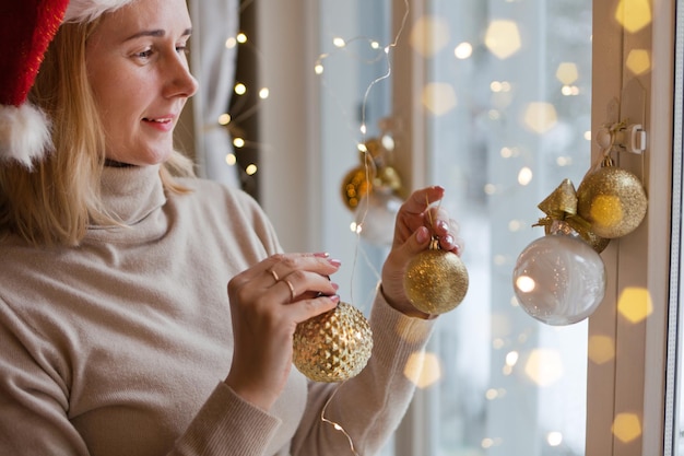 Woman decorating window using golden christmas balls and lights bokeh blond wearing red santa hat