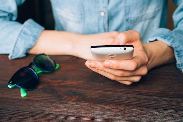 Woman in a denim shirt holds a mobile phone and sitting at a wooden table on which lie sunglasses