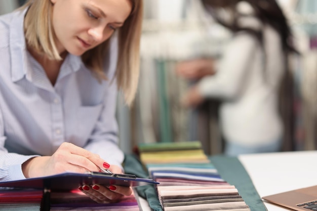 Woman designer writing with pen on clipboard in curtain salon closeup online ordering of design