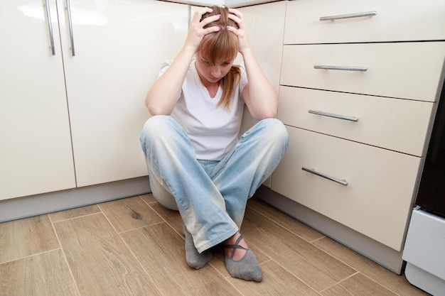 A woman in despair sits on the floor in the kitchen and holds her head