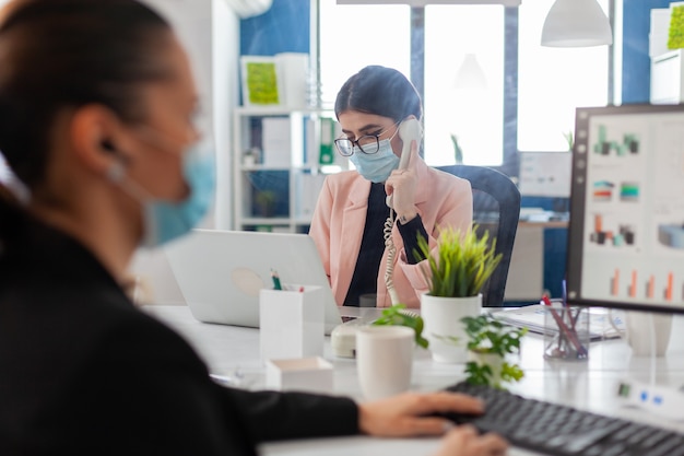 Woman discussing on phone in new normal office wearing face mask as safety prevention behind plastic shield, keeping social distancing with coworker during coronavirus flu outbreak.