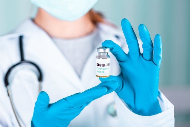 Woman doctor or nurse in uniform and gloves wearing face mask protective in lab, holding medicine vial vaccine bottle with COVID-19 Coronovirus vaccine label