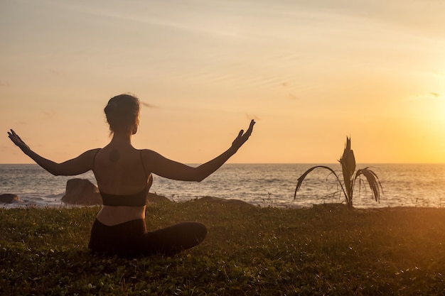 Woman does yoga position arms raised on tropical sea coast or ocean beach outdoors at sunset