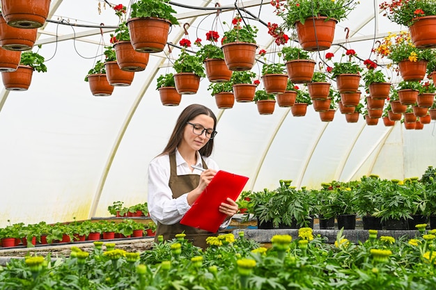 Woman doing list of plants Portrait of a young woman at work in greenhouse in uniform and clipboard in her hand