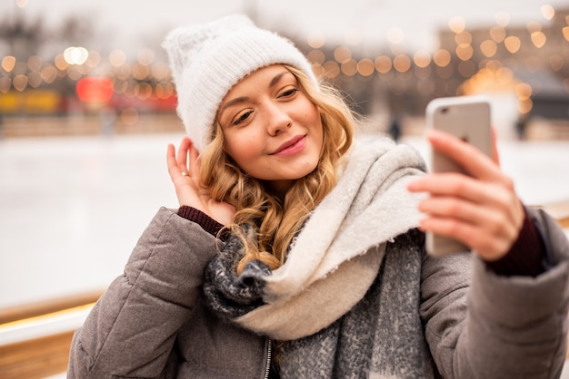 Woman doing selfie on the background of festive Christmas ice rank. Lady wearing winter warm knitted clothes.