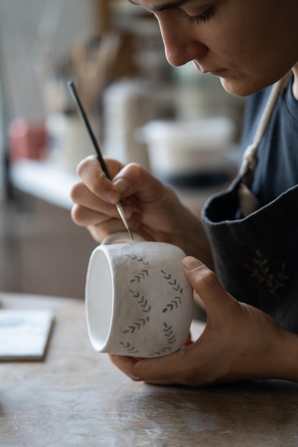 Woman draws pattern with dark paint on white ceramic mug using brush on blurred background