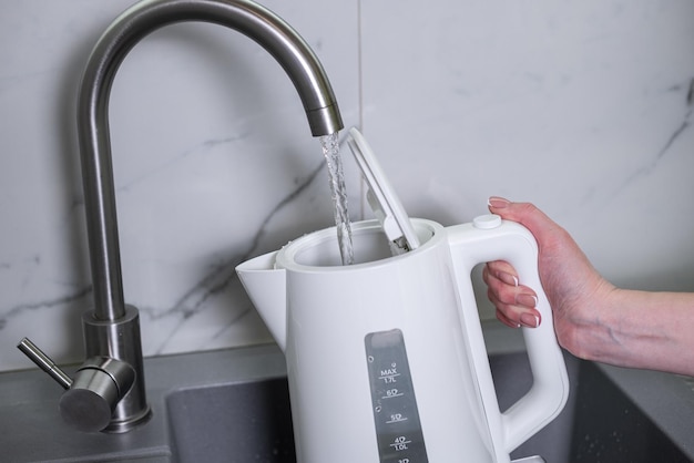 A woman draws water into an electric kettle in the background of the kitchen It's time for breakfast and tea Modern electric kettle Kettle for boiling water