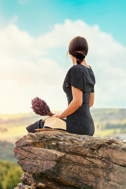 Woman in a dress and a hat with a lavender bouquet in her hands on a hill in summer sunset