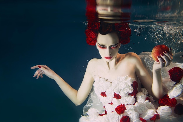 Woman in dress made of white and red roses underwater. Fairy tale, art, fashion concept