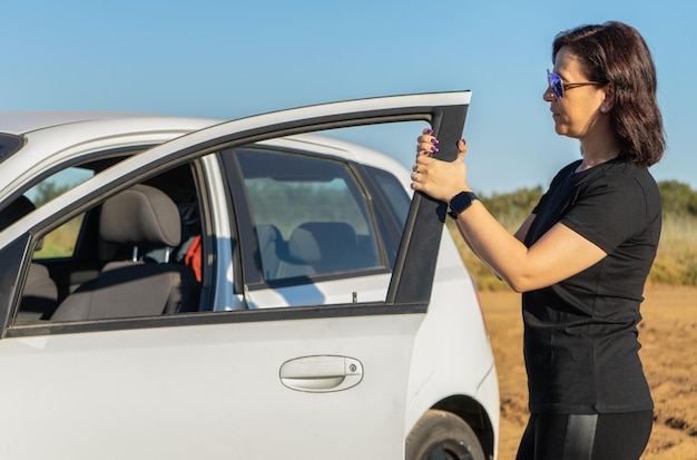 Woman dressed in black holding the door of a white car wearing sunglasses with the sun on her face