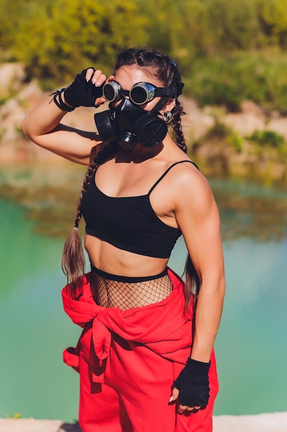 Woman dressed in a steampunk suit in a defiant attitude with her arms crossed respirator on the face against the background of a dirty lake