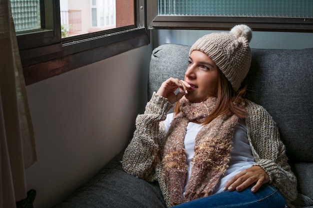 Woman dressed in warm clothes because of the cold winter, sitting on her sofa next to the window. 